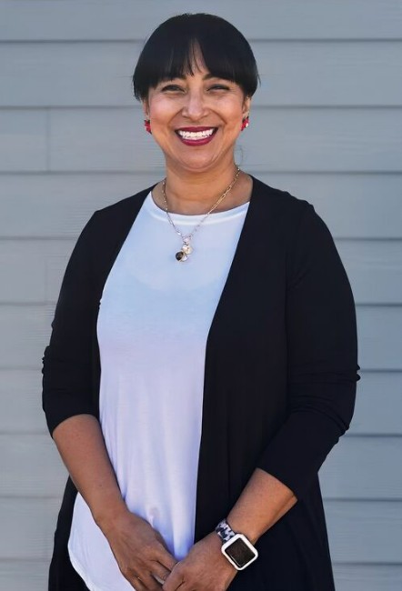 Meet The Team: A woman with dark hair and bangs, wearing a white top, black cardigan, necklace, and smartwatch, smiles while standing in front of a gray wall.