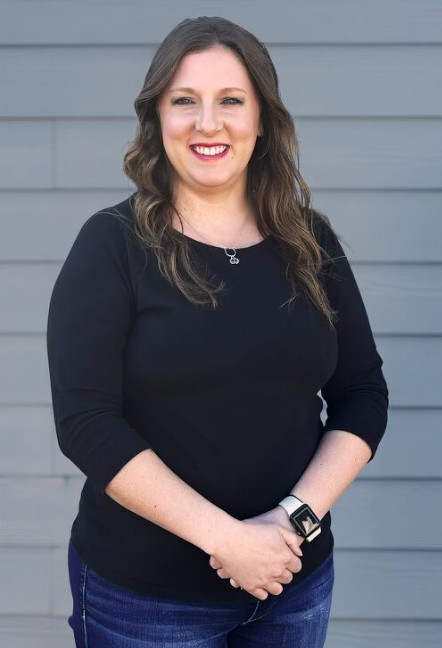 Meet the Team: A woman with long brown hair wearing a black top and jeans stands in front of a gray wall, smiling with her hands clasped.