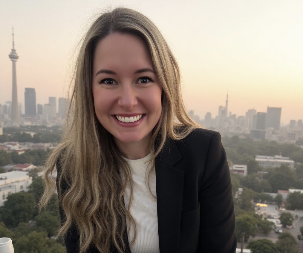 Meet The Team: A woman with long blonde hair smiles at the camera, with a city skyline and tower visible in the background during sunset.